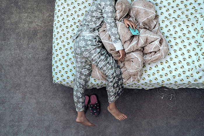 Person in floral pajamas on bed, holding a phone, embracing traditional rest values from older generations.