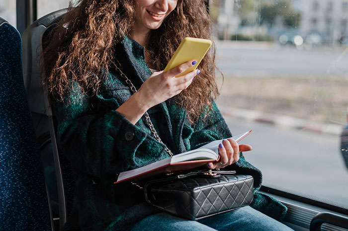 Woman on a bus using a smartphone and smiling, with a notebook in her lap, embracing boomer habits.