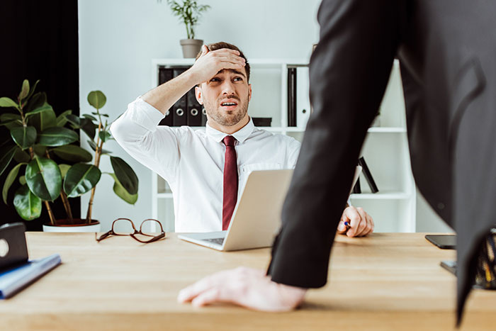 Employee looking stressed at a desk, next to a laptop, as a manager stands over them in an office setting. Employee looking stressed at a desk, next to a laptop, as a manager stands over them in an office setting.