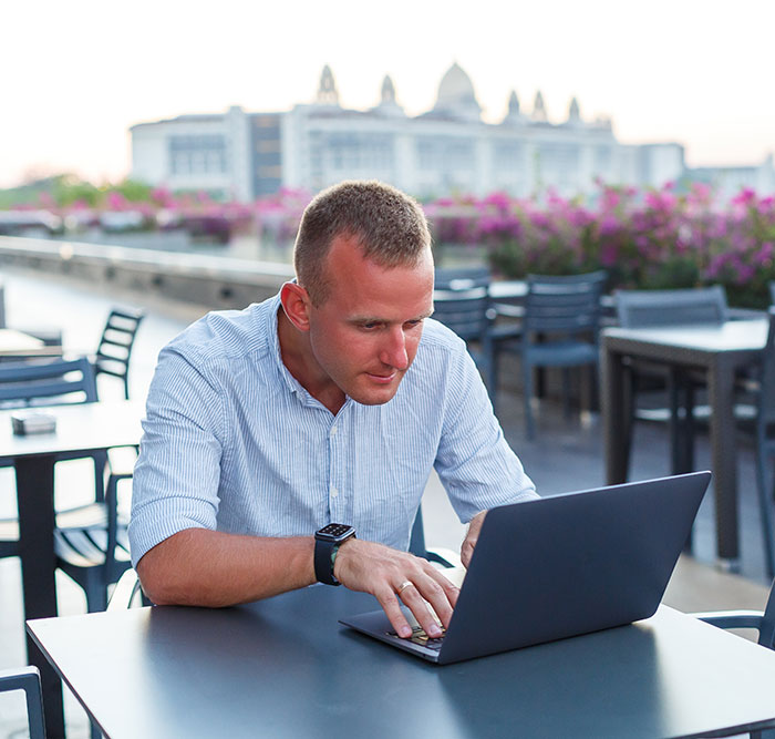 Man in blue shirt focusing on laptop while seated outdoors at a café, representing remote work challenges faced on vacation. Man in blue shirt focusing on laptop while seated outdoors at a café, representing remote work challenges faced on vacation.