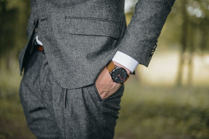 A person in a gray suit standing outdoors, hand in pocket, wearing a stylish watch, symbolizing professional attire.