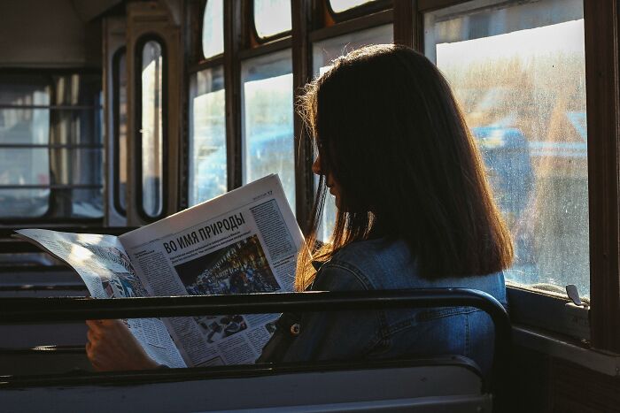 A woman reading a newspaper on a sunlit bus, a normal sight in society.