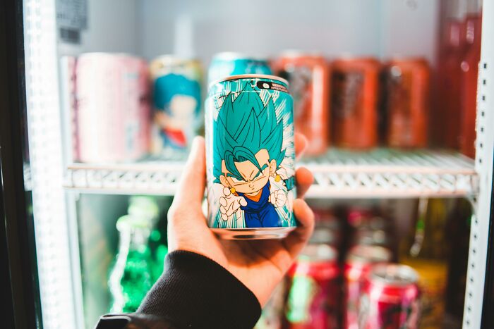 Hand holding a colorful anime-themed soda can in front of a fridge, showcasing small-thing-middle-class-home.