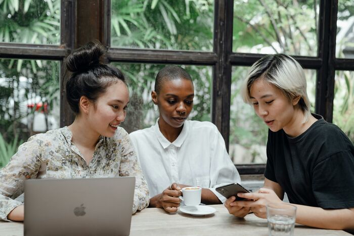 Three adults at a café table with a laptop and coffee, sharing experiences and looking at a phone together.