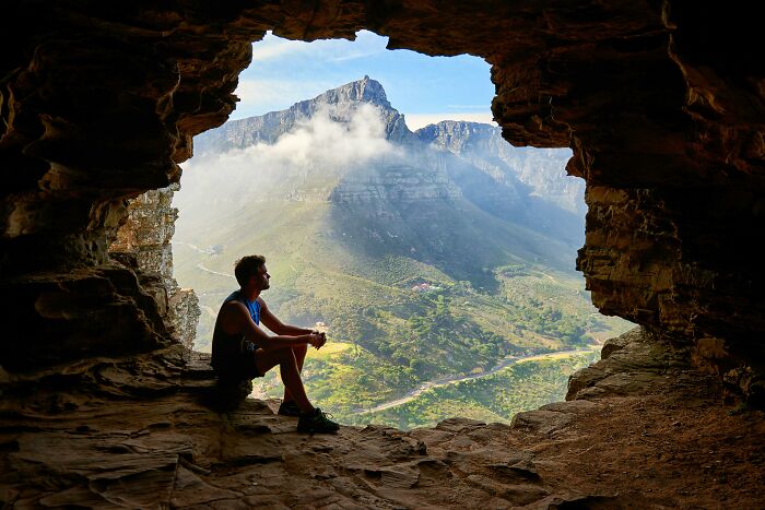 Man sitting in a cave, overlooking a breathtaking natural wonder landscape with mountains and clouds in the distance. Man sitting in a cave, overlooking a breathtaking natural wonder landscape with mountains and clouds in the distance.