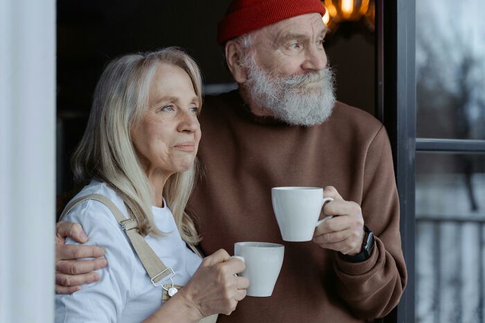 Elderly couple in casual attire holding mugs by a window, embracing society's normal.