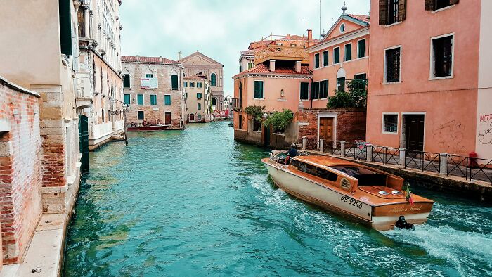 A boat navigates a canal in a touristy area, lined with colorful buildings under a cloudy sky.