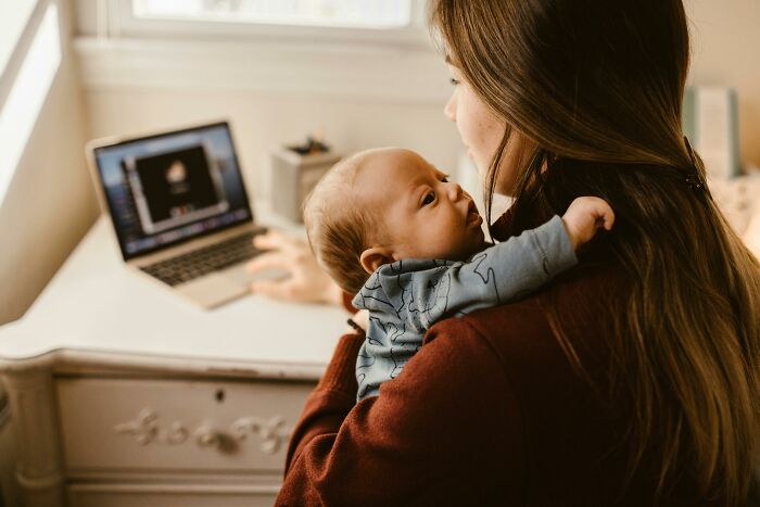 A woman holding a baby while working on a laptop, challenging common stereotypes.