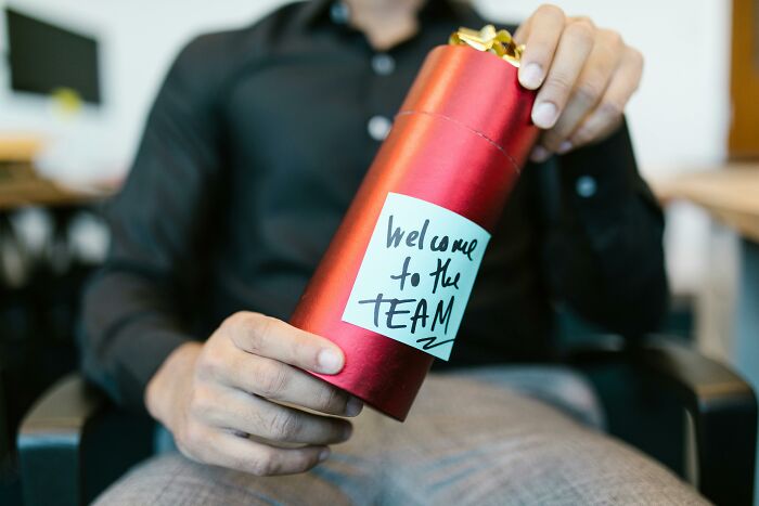 Person holding a red gift tube with "Welcome to the TEAM" note, dressed casually in an office setting.
