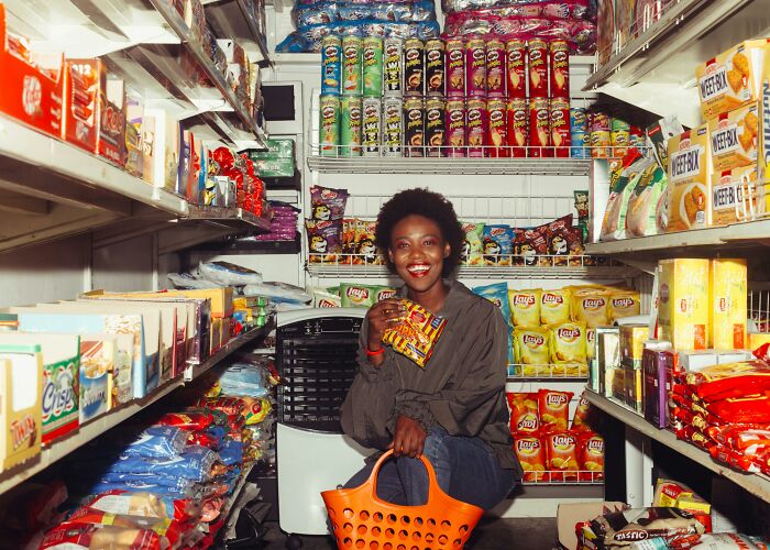 Woman shopping with basket, surrounded by snacks in a small middle-class home store aisle.