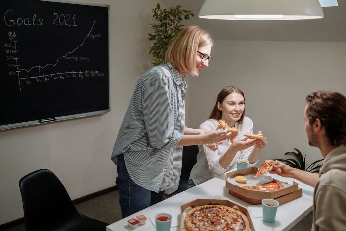 Three adults enjoying pizza in an office with a graph on a chalkboard, symbolizing adult experiences.