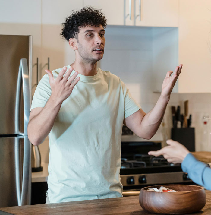 Man discussing in kitchen while gesturing, partner's hand pointing back, highlighting relationship and sterilization topic. Man discussing in kitchen while gesturing, partner's hand pointing back, highlighting relationship and sterilization topic.