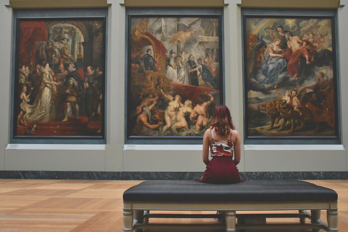A woman sitting on a bench, observing three large paintings, contemplating real vs. forgery in an art gallery. A woman sitting on a bench, observing three large paintings, contemplating real vs. forgery in an art gallery.