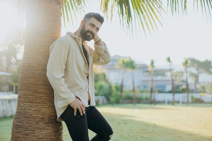 Man with beard leaning on palm tree in sunny park, exemplifying pretty privilege.