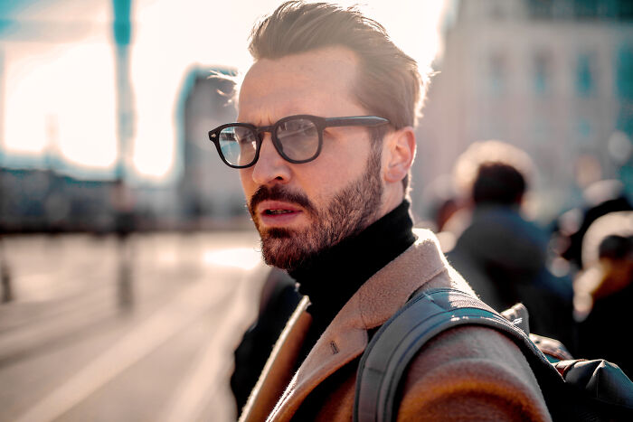 Man with glasses and beard wearing a coat, representing pretty privilege in a city setting.