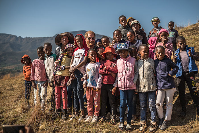 A man in a Harvard shirt surrounded by smiling children outdoors in a mountainous area, related to charity and controversy. A man in a Harvard shirt surrounded by smiling children outdoors in a mountainous area, related to charity and controversy.