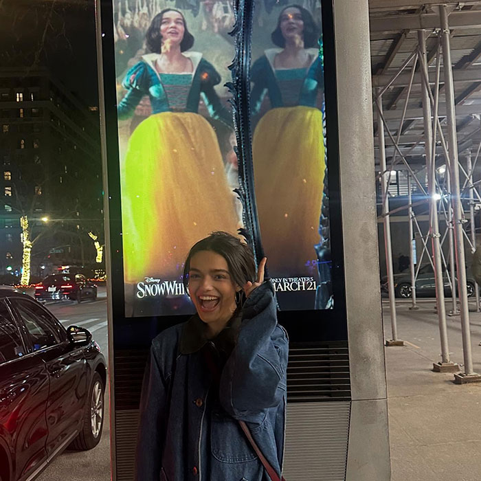 A woman smiles beside a digital poster for Disney's 'Snow White' at night, with city lights in the background. A woman smiles beside a digital poster for Disney's 'Snow White' at night, with city lights in the background.