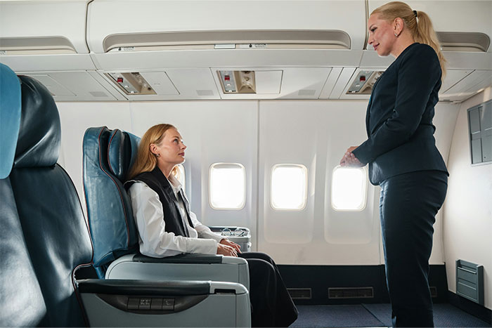 Passenger sits in airplane seat while flight attendant stands nearby discussing seat arrangements. Passenger sits in airplane seat while flight attendant stands nearby discussing seat arrangements.