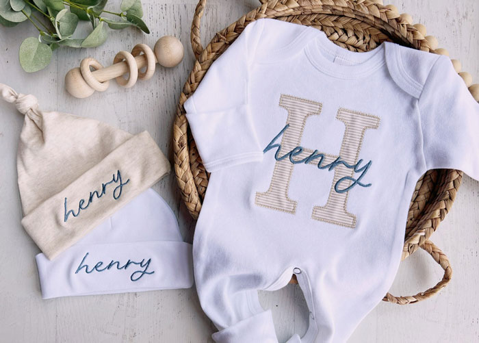 Baby clothes with "Henry" name, breaking tradition, on a woven basket with a wooden toy and greenery nearby. Baby clothes with "Henry" name, breaking tradition, on a woven basket with a wooden toy and greenery nearby.