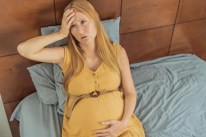 Pregnant woman in a yellow dress, looking concerned while sitting on a bed, contemplating family name tradition. Pregnant woman in a yellow dress, looking concerned while sitting on a bed, contemplating family name tradition.