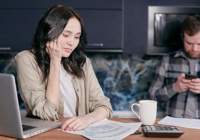 Woman concerned over college fund papers at kitchen table, man using phone in background. Woman concerned over college fund papers at kitchen table, man using phone in background.