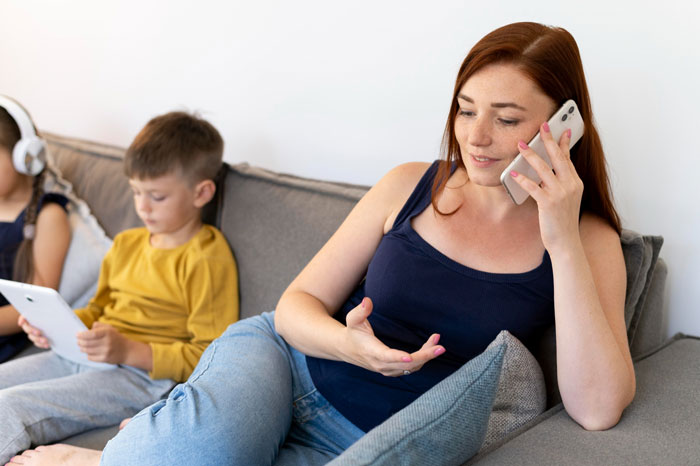 Woman on phone while kids play with tablets on couch, parenting theme.