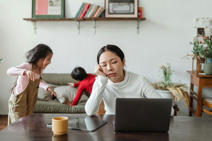 A tired woman at a table with a laptop, while two kids play in the background.