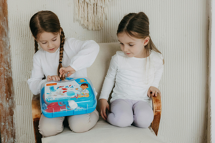Two young girls sitting on a chair, one opening a blue toy medical kit, reflecting sibling dynamics. Two young girls sitting on a chair, one opening a blue toy medical kit, reflecting sibling dynamics.