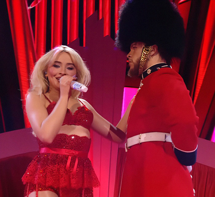 Singer performing in red outfit with guard in themed concert scene. Singer performing in red outfit with guard in themed concert scene.