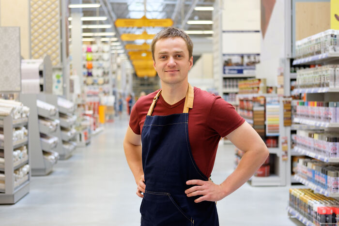 Man in apron smiling confidently in a store aisle, symbolizing routine yet stuck in insignificant tasks forever.