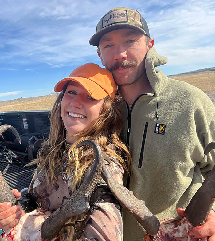 A smiling couple outdoors, posing with large antlers, against a rural landscape backdrop. A smiling couple outdoors, posing with large antlers, against a rural landscape backdrop.