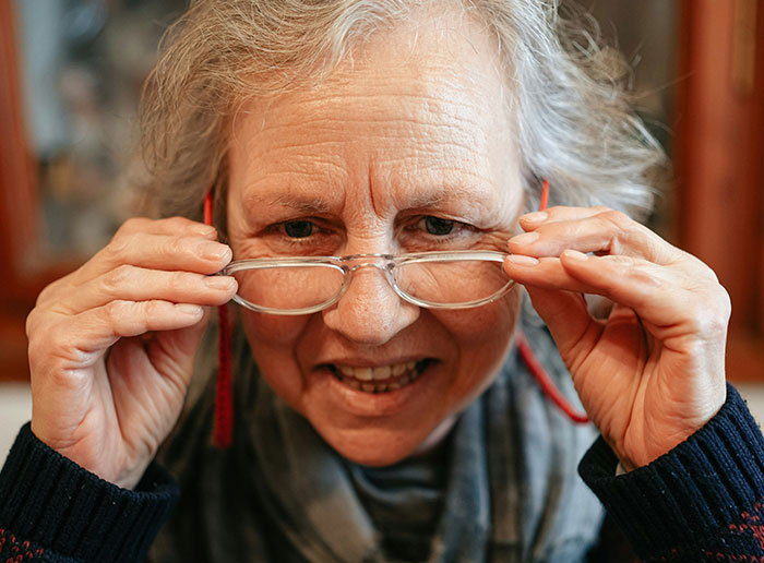 Elderly woman adjusting glasses, smiling and leaning forward, inside a cozy setting.