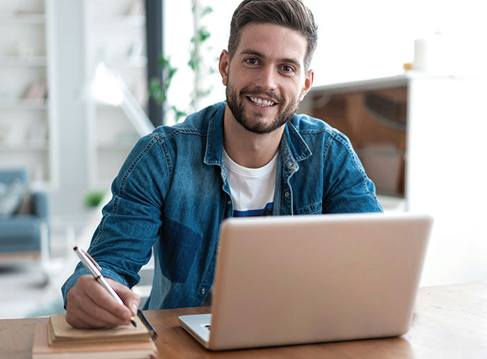 Man smiling at laptop, wearing a denim shirt, taking notes about bosses from hell stories.