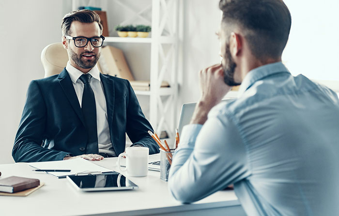 Two men in a business meeting, one wearing glasses, discussing work across a desk in a modern office setting.