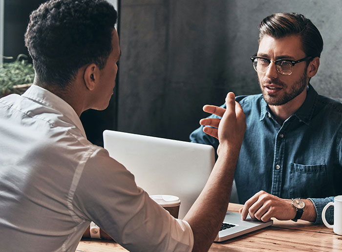 Two men discussing at a table, one wearing glasses and denim, possibly talking about difficult bosses.