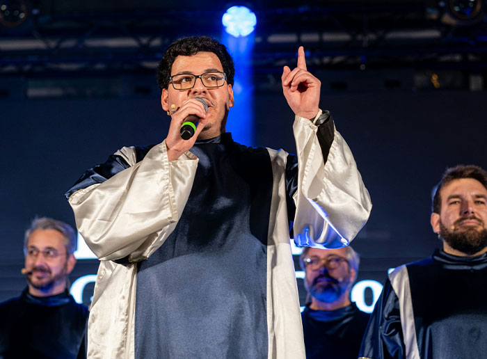 Man on stage with microphone, pointing up, surrounded by others. Event setting.