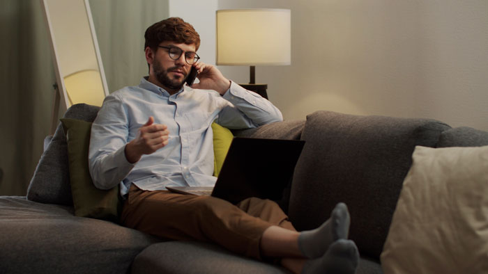A gay man, flabbergasted, sits on a couch with a laptop, talking on the phone about being excluded from a will. A gay man, flabbergasted, sits on a couch with a laptop, talking on the phone about being excluded from a will.