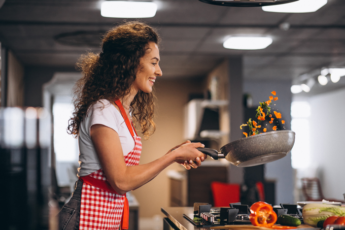 Woman cooking with skillet in modern kitchen, wearing a red apron, smiling while tossing vegetables in the air. Woman cooking with skillet in modern kitchen, wearing a red apron, smiling while tossing vegetables in the air.