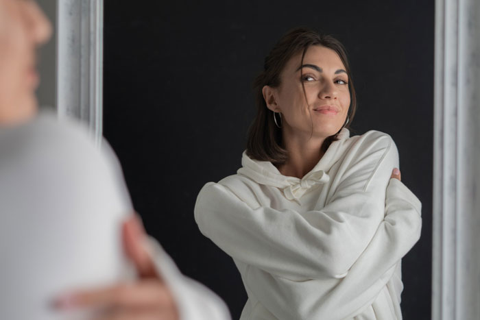 Woman in a white hoodie smiling in front of a mirror, crossing her arms confidently, depicting a selfish girlfriend theme. Woman in a white hoodie smiling in front of a mirror, crossing her arms confidently, depicting a selfish girlfriend theme.