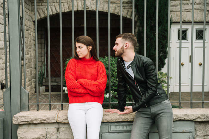 Man and woman standing outside by a fence, man talking while woman in red sweater crosses her arms, looking away. Man and woman standing outside by a fence, man talking while woman in red sweater crosses her arms, looking away.