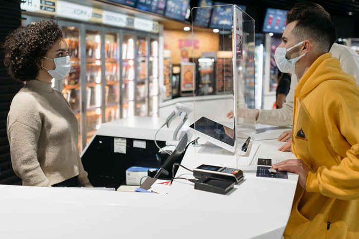 Moviegoer at a concession stand, wearing a mask and yellow hoodie, speaking to a staff member during a transaction. Moviegoer at a concession stand, wearing a mask and yellow hoodie, speaking to a staff member during a transaction.