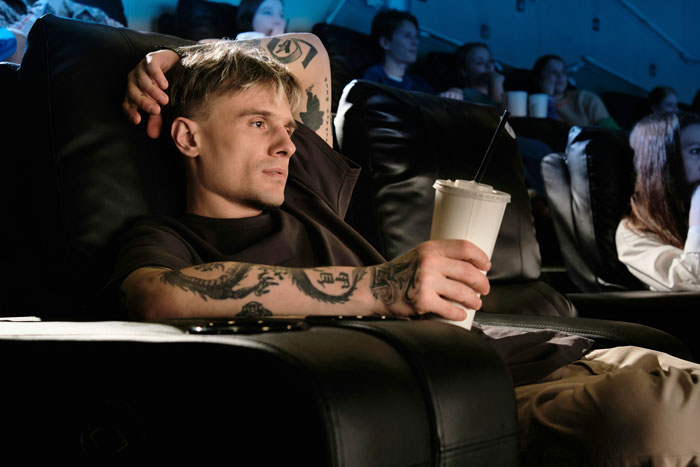 Moviegoer with tattoos and drink, relaxed in a theater, unaware of upcoming spoilers after cutting in line. Moviegoer with tattoos and drink, relaxed in a theater, unaware of upcoming spoilers after cutting in line.
