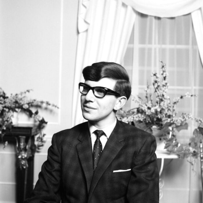 Young man in a suit and glasses during the JFK era, seated in a classic interior setting with floral decor.