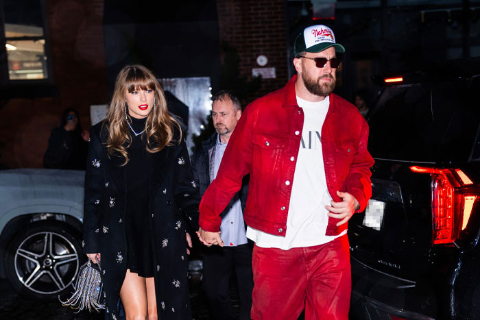 Taylor Swift and Travis Kelce holding hands in a city street, she in a black outfit and he in a red jacket and cap, exits a black SUV.