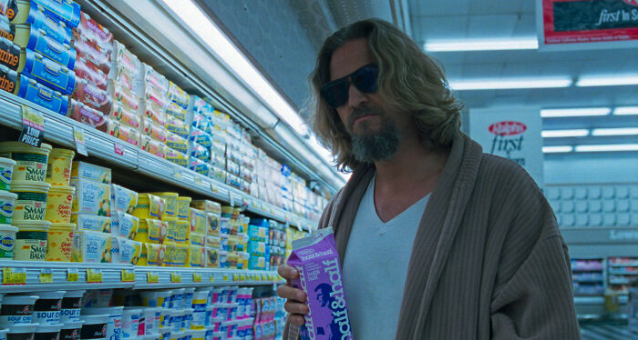 Actor in a grocery store aisle holding half-and-half milk, wearing sunglasses and a casual robe.