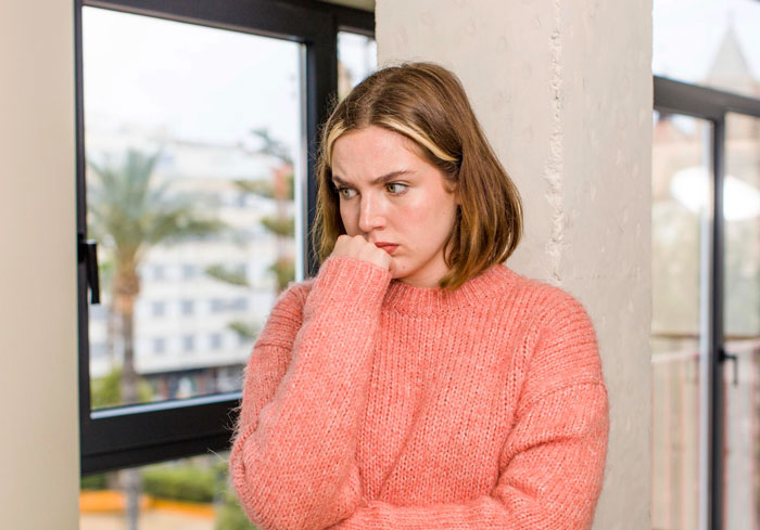 A woman in a pink sweater looks upset, standing by a window, reflecting on exclusion from a birthday party. A woman in a pink sweater looks upset, standing by a window, reflecting on exclusion from a birthday party.