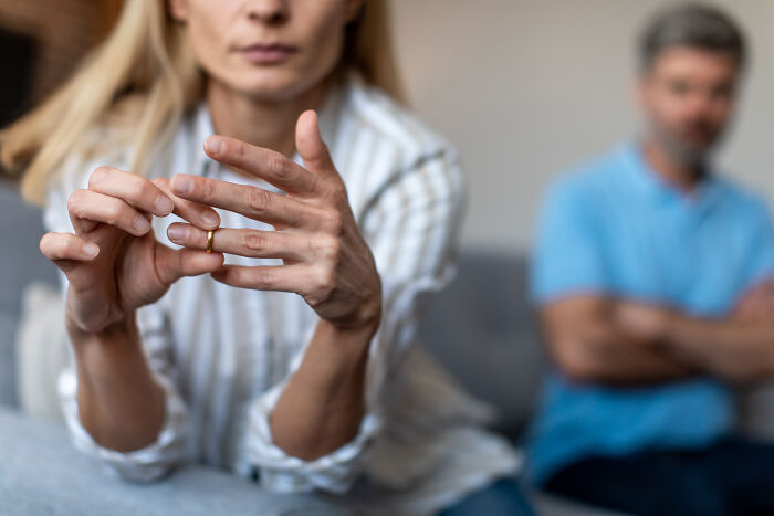 Woman removing ring, sitting on a couch with a blurred man in the background, symbolizing unexpected things in reality.