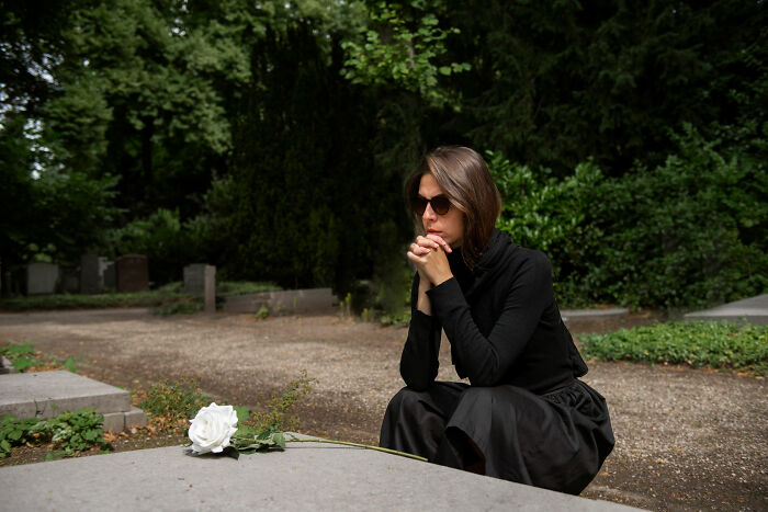 A woman in dark clothing kneels by a grave, deep in thought, in a serene cemetery setting, symbolizing unexpected reality.