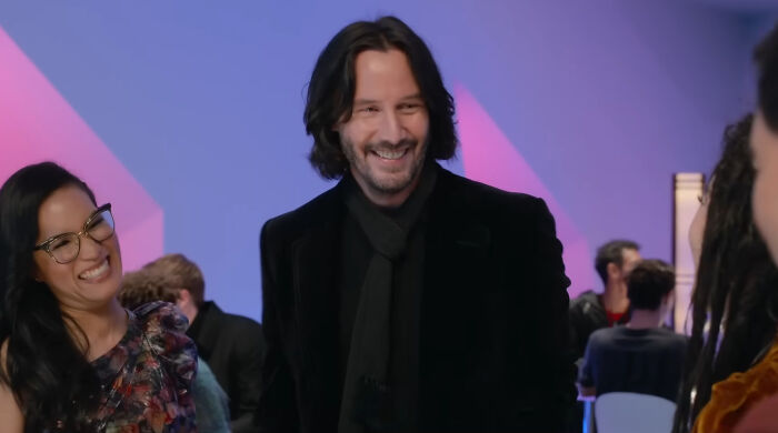 Smiling celebrity at an event, dressed in black, surrounded by people against a colorful background.