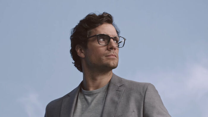 A celebrity in glasses and a gray suit poses against a clear sky, capturing attention and admiration online.